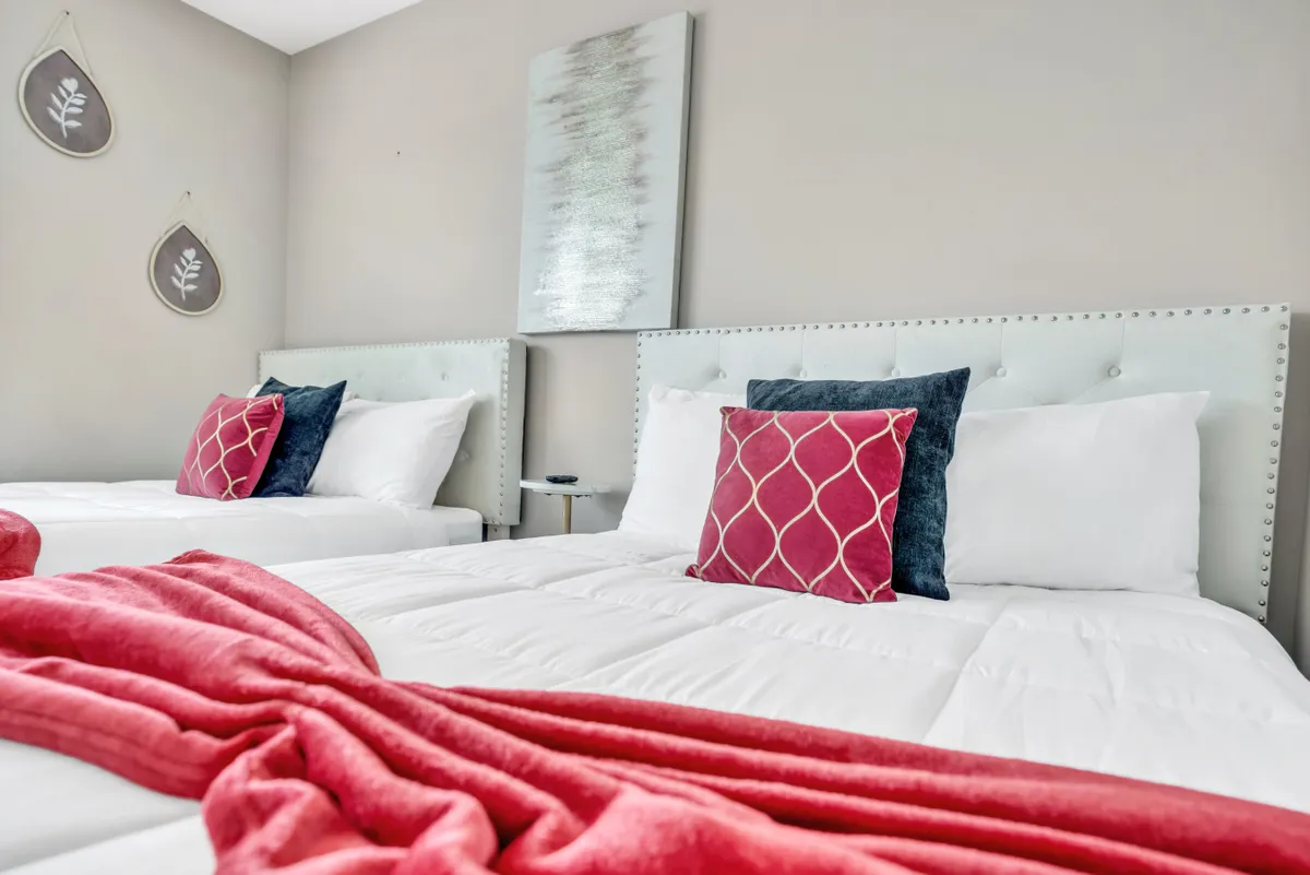 A cozy bedroom with two beds adorned with decorative pillows and a plush blanket, complemented by modern wall art. The room exudes warmth and comfort, perfect for families or guests seeking a restful environment.