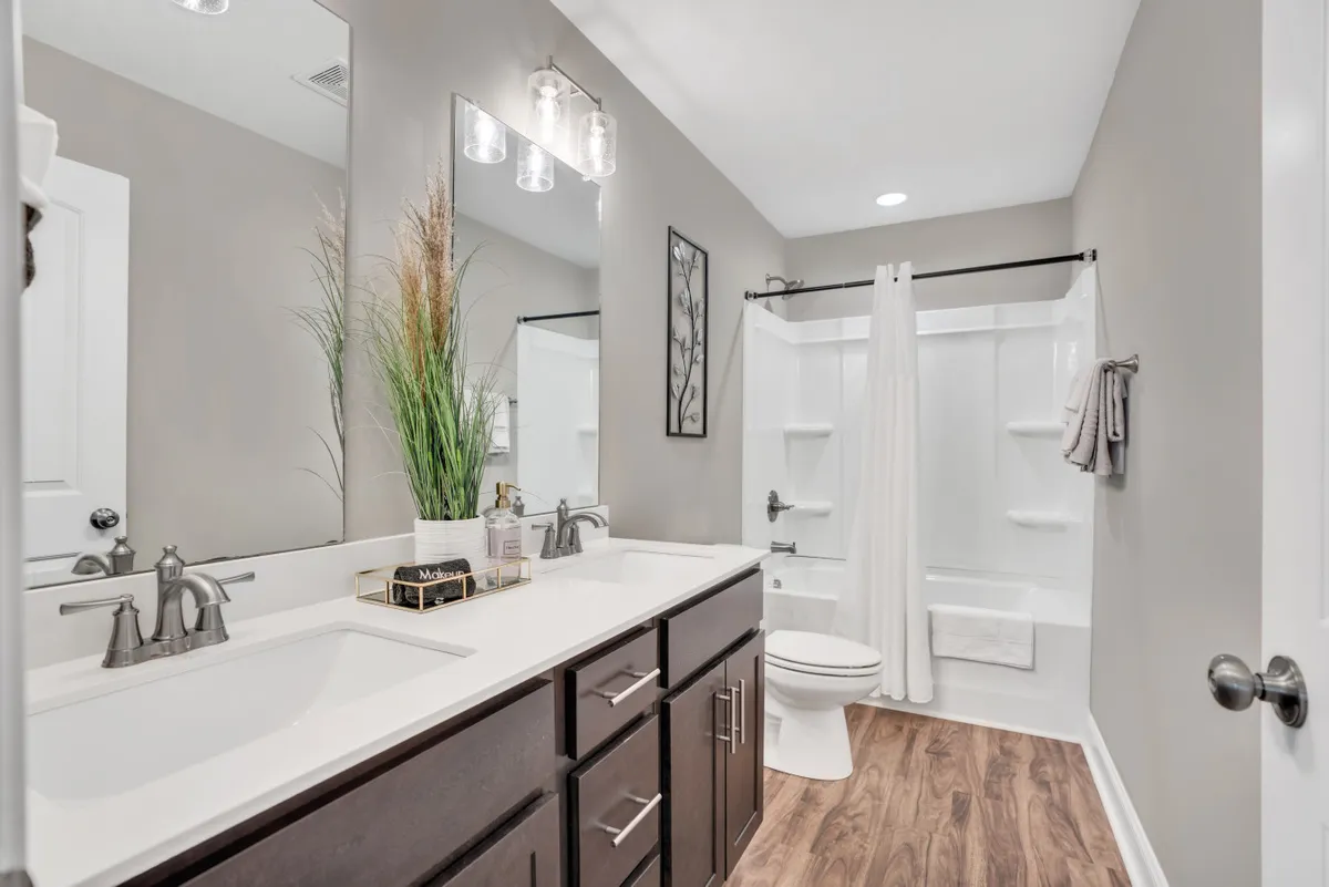 A modern bathroom with a double sink, large mirror, decorative plant, and framed wall art. The space feels calm and inviting, offering an ideal spot for personal care and relaxation.