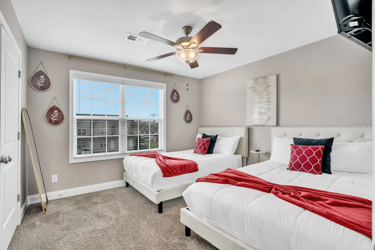 This image depicts a bright bedroom with two beds dressed in crisp white linens and vibrant red accents. The beds are positioned near a large window that lets in abundant natural light, and the space is decorated with tasteful wall hangings. The overall setting appears inviting and spacious, making it ideal for families or groups seeking comfort and relaxation close to Fort Campbell.