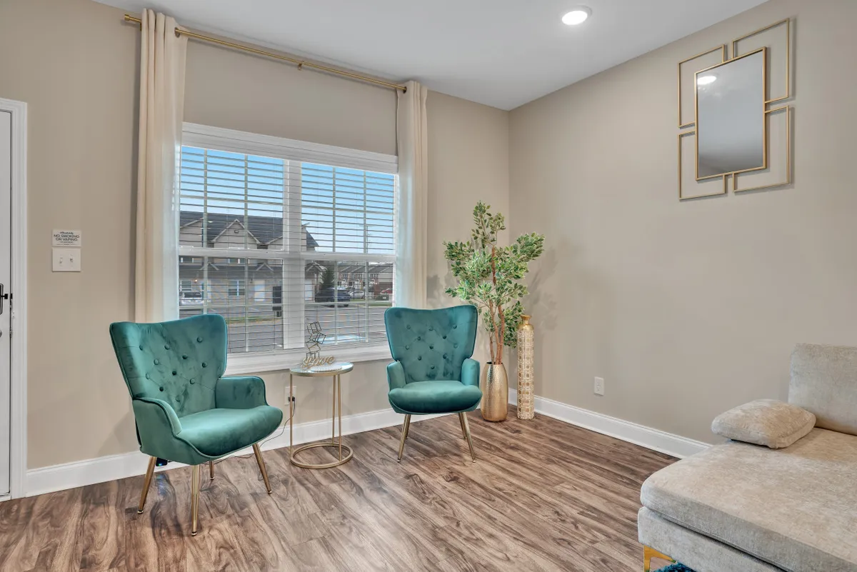 A bright, inviting entryway or sitting nook featuring two turquoise chairs, a small round side table, a decorative plant, and a window with blinds. Natural light floods the space, creating a relaxing spot for reading or conversation.
