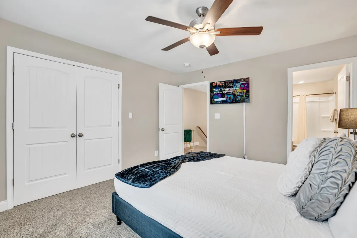 This image presents a tranquil bedroom setup featuring a bed with decorative pillows, a ceiling fan for comfort, a wall-mounted television for relaxation, and double doors leading to a closet. The room exudes a sense of coziness and offers a restful retreat for guests.